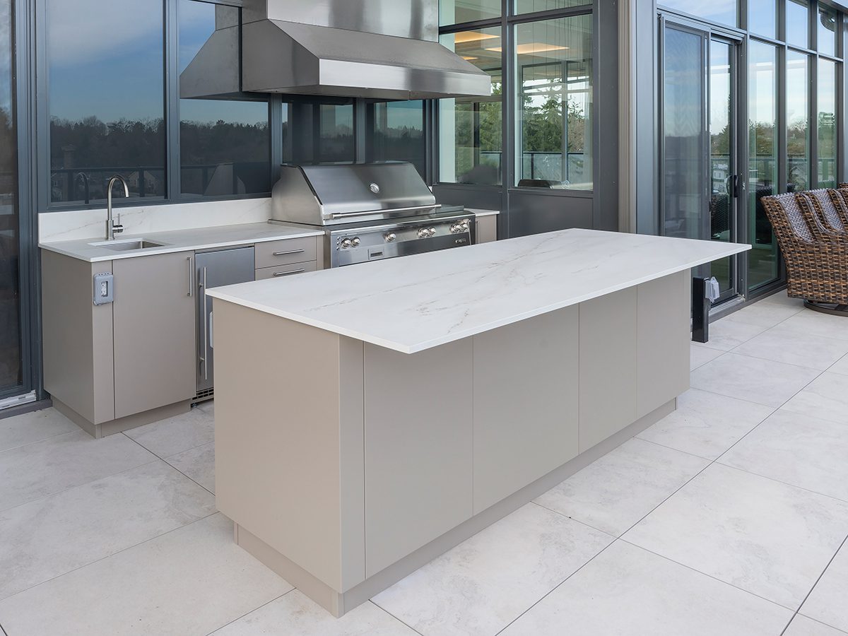Modern galley outdoor kitchen on a rooftop terrace featuring taupe cabinetry, stainless grill with vent hood, sink, and a quartz island worktop