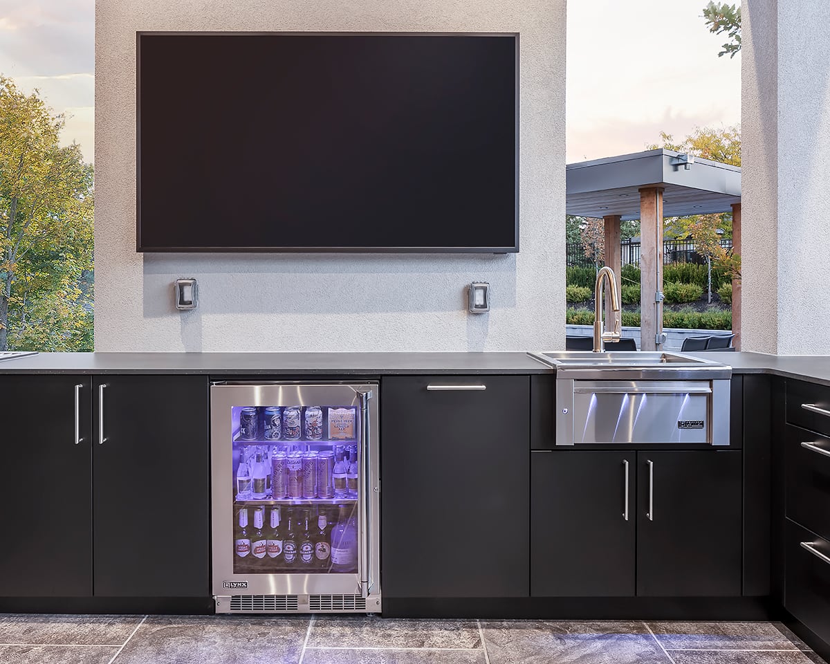 Outdoor kitchen beverage station with glass-door fridge, bar sink, and wall-mounted TV in matte-black cabinetry