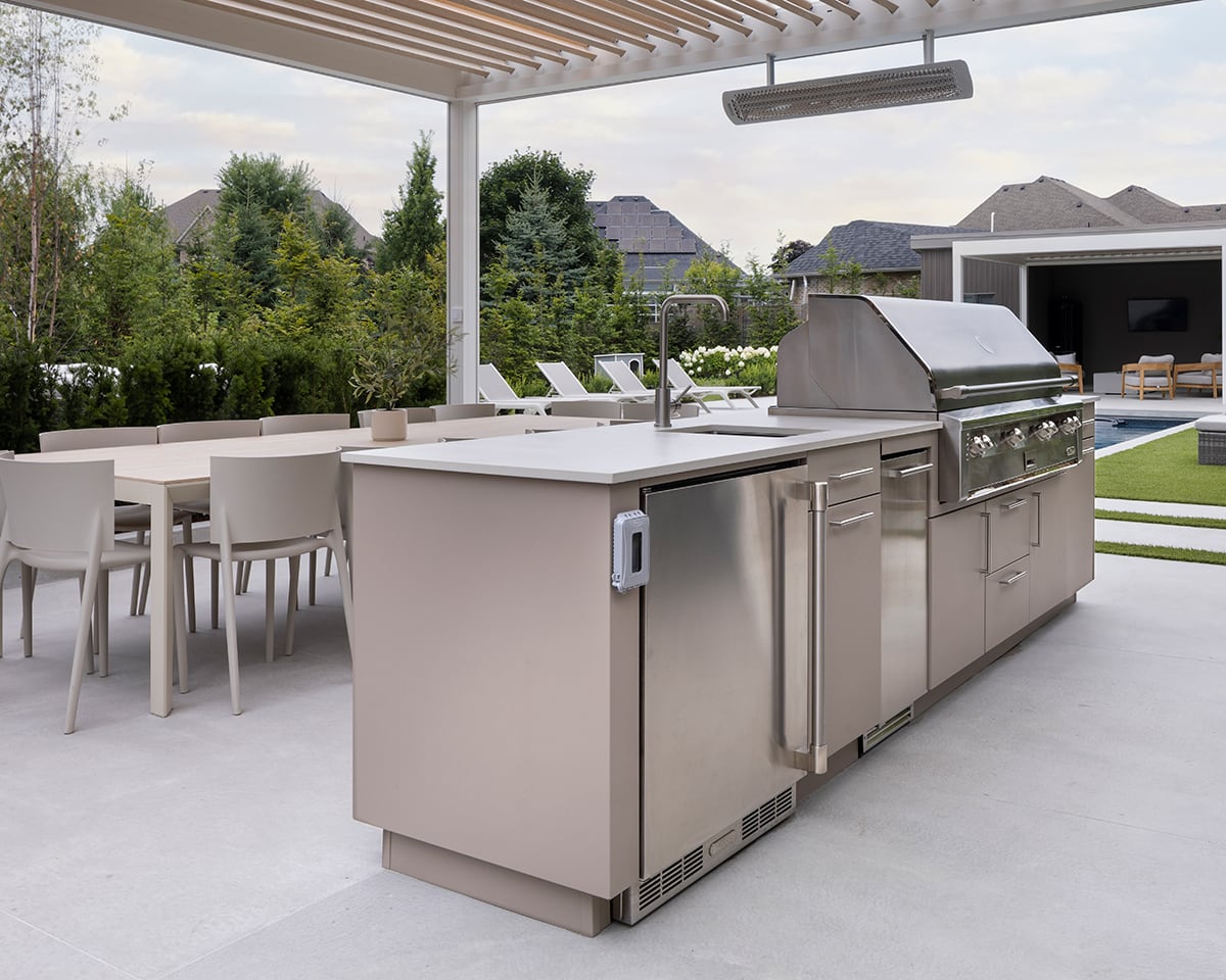 Pergola-covered island kitchen with stainless grill, fridge, and modern dining table beside a pool in a landscaped backyard