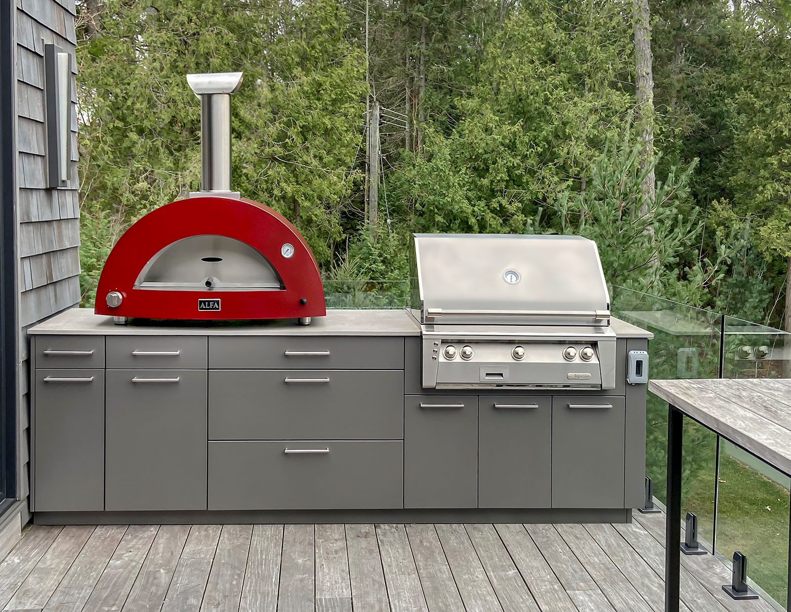 Straight-line outdoor kitchen on cottage deck featuring red Alfa pizza oven, built-in stainless grill, and charcoal-grey weatherproof cabinetry