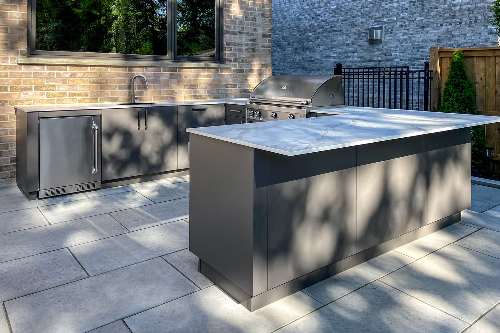 U-shaped outdoor kitchen with charcoal-gray cabinets, stainless grill and fridge, and marble-look quartz countertop on a stone patio