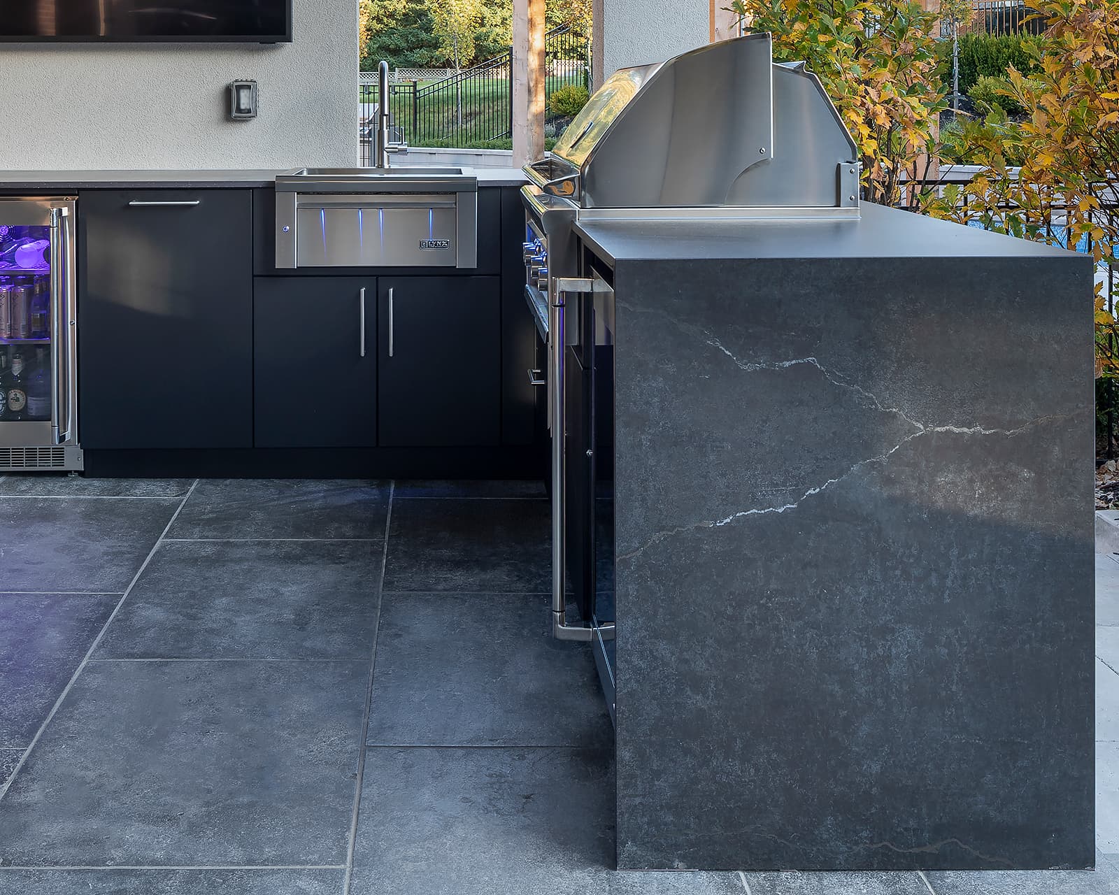 Close-up of a dark Dekton porcelain waterfall countertop edge on an outdoor kitchen in Toronto, showing seamless miter joint with continuous veining beside a stainless Lynx grill and matte black aluminium cabinets.