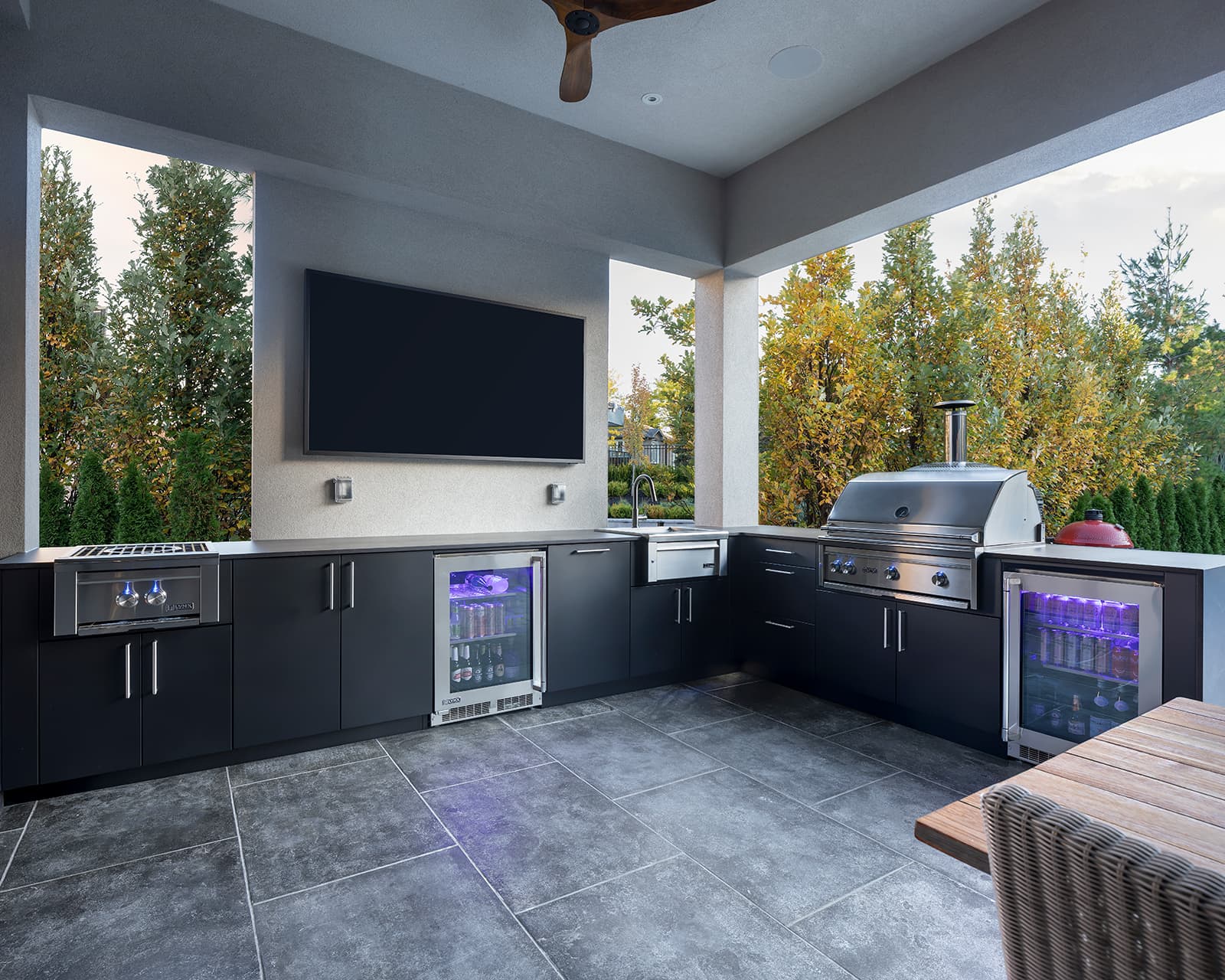 Wide-angle view of a covered L-shaped outdoor kitchen in Toronto featuring a Lynx power burner, 36-inch stainless grill, dual illuminated beverage fridges, 24-inch sink, matte black aluminium cabinetry, and wall-mounted TV.