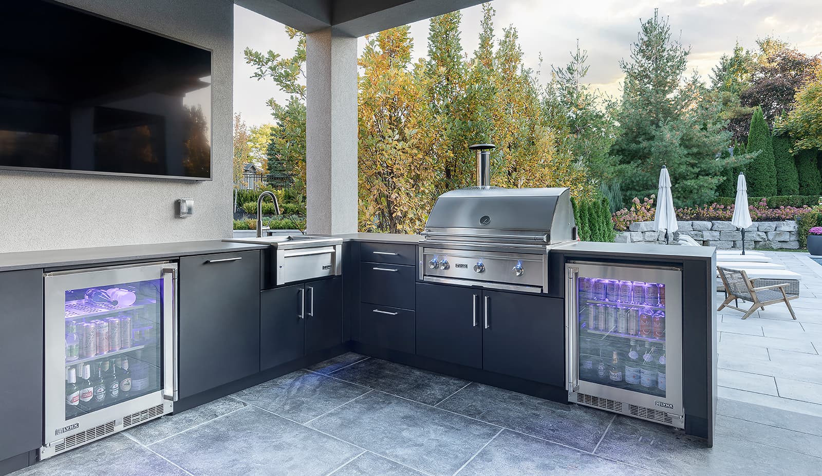 Close-up of a modern L-shaped outdoor kitchen in Toronto showcasing a 36-inch Lynx Professional stainless-steel grill, twin glass-door beverage fridges, farmhouse sink, and porcelain countertops set within matte black aluminium cabinetry under a covered patio.