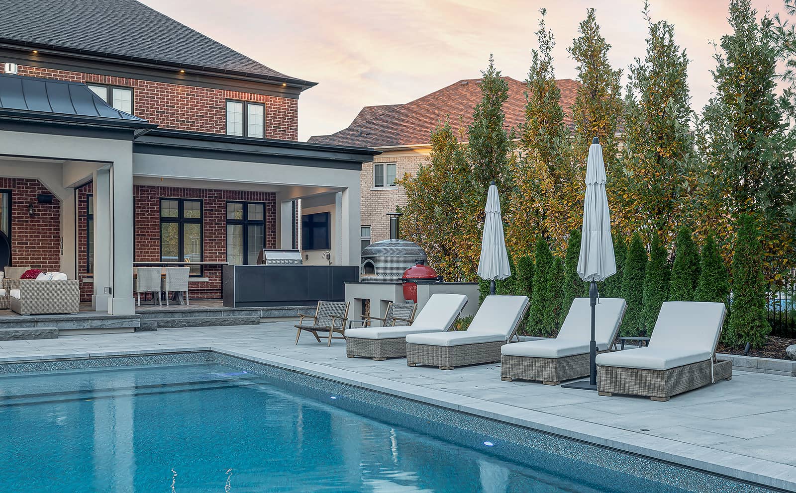 Wide view of a Toronto backyard showing a poolside L-shaped outdoor kitchen with a Lynx stainless-steel grill and pizza oven under a cedar-clad porch, aligned with lounge chairs and dining area for seamless entertaining.
