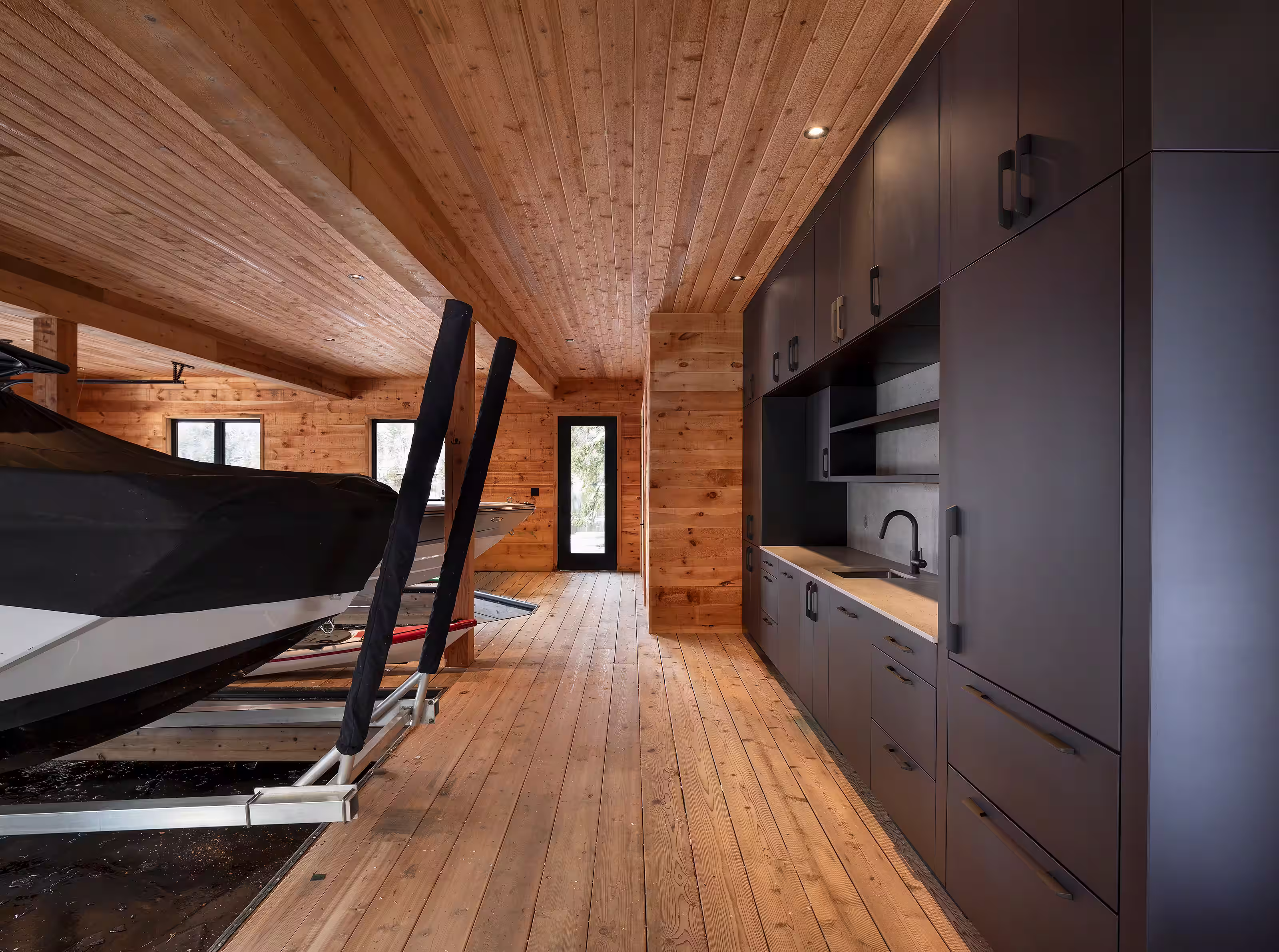 Modern Muskoka boathouse interior featuring a black and white marble waterfall island, minimalist greige cabinetry, and a large glass garage door opening to a wooded shoreline.