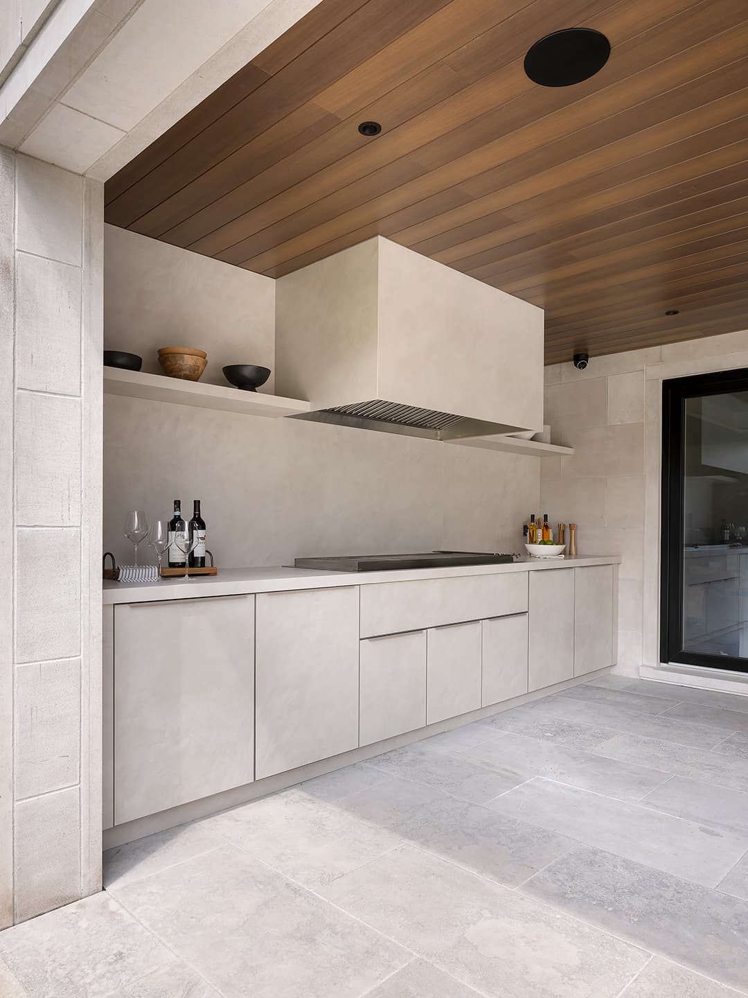 Angled view of a covered modern outdoor kitchen featuring limestone cabinetry a built in grill and warm wood ceiling