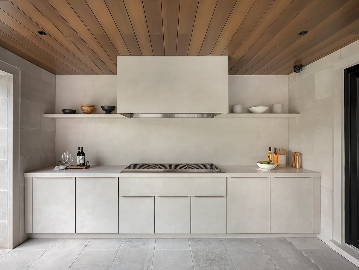 Front view of a covered modern outdoor kitchen featuring limestone cabinetry a built in grill and warm wood ceiling