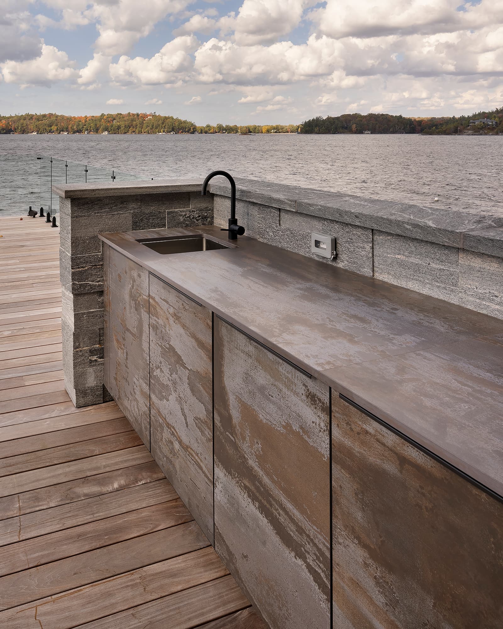 Outdoor kitchen cabinetry with an integrated sink on a Muskoka boathouse deck, featuring weathered metal finishes, stone surround, and open lake views.