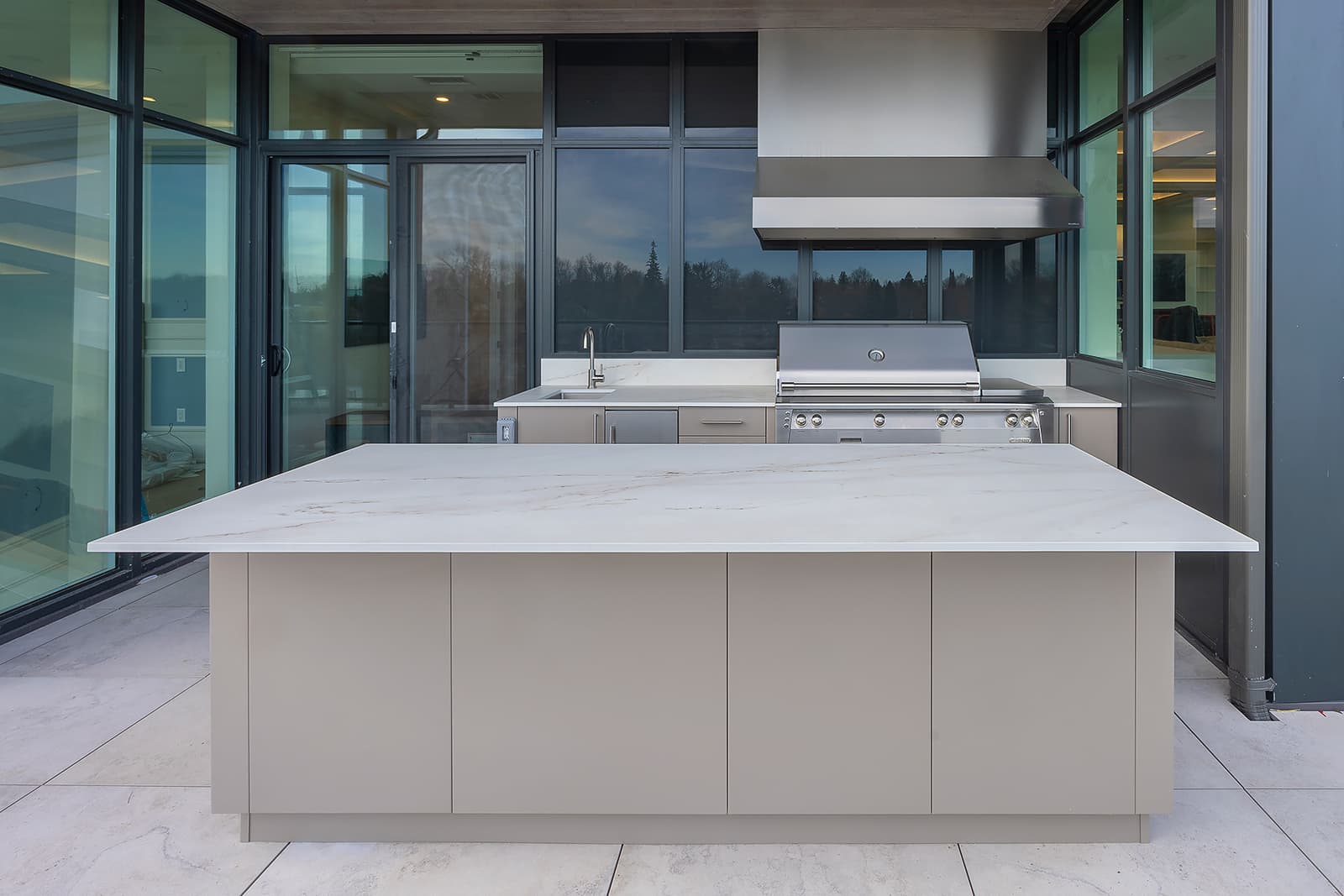 Modern outdoor kitchen with stone island, handleless cabinets, stainless grill & sink framed by glass windows and tiled floor.